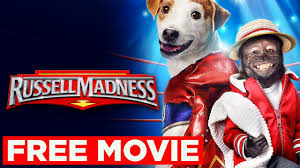 A energetic Jack Russell Terrier named Russell wears a tiny wrestling championship belt around his waist, standing triumphantly on a wrestling ring turnbuckle with one paw raised in victory. Behind him, a colorful wrestling arena crowd cheers, holding signs that read "Go Russell!" and "The Fury Rules!" Two larger dogs (a bulldog and a German Shepherd) in opposing corners snarl playfully, wearing wrestling singlets. The scene has bright, vibrant lighting like a real WWE event, with dramatic spotlights highlighting the furry competitors.
